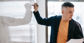 Man drawing on whiteboard holding paperwork and looking to right